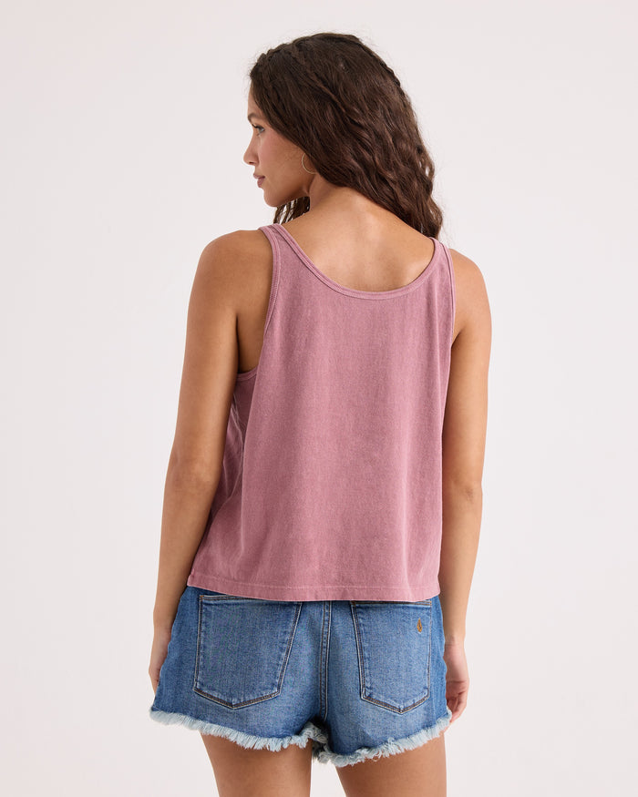 A woman with wavy brown hair wears the Volcom Women's Amakua Bank Tank in mauve paired with blue denim shorts. She is standing with her back to the camera against a plain light background.