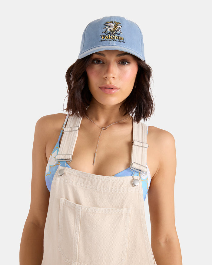 A person wearing a Volcom Women's Ameristone Hat in horizon blue, beige overalls, a blue patterned top, and a delicate necklace stands against a plain white background.