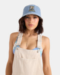 A person wearing a Volcom Women's Ameristone Hat in horizon blue, beige overalls, a blue patterned top, and a delicate necklace stands against a plain white background.