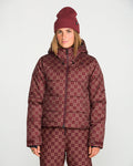 A woman stands against a plain white background wearing a matching maroon beanie, Volcom Women’s Avalon Puff Jacket with recycled insulation, and checkerboard patterned pants.