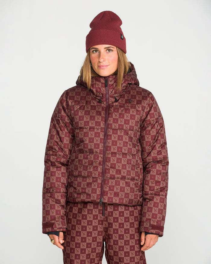 A woman stands against a plain white background wearing a matching maroon beanie, Volcom Women’s Avalon Puff Jacket with recycled insulation, and checkerboard patterned pants.