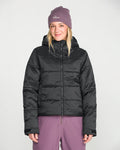 A person wearing a purple beanie, purple pants, and the Volcom Women’s Avalon Puff Jacket with recycled insulation stands facing forward against a plain white background, smiling slightly.
