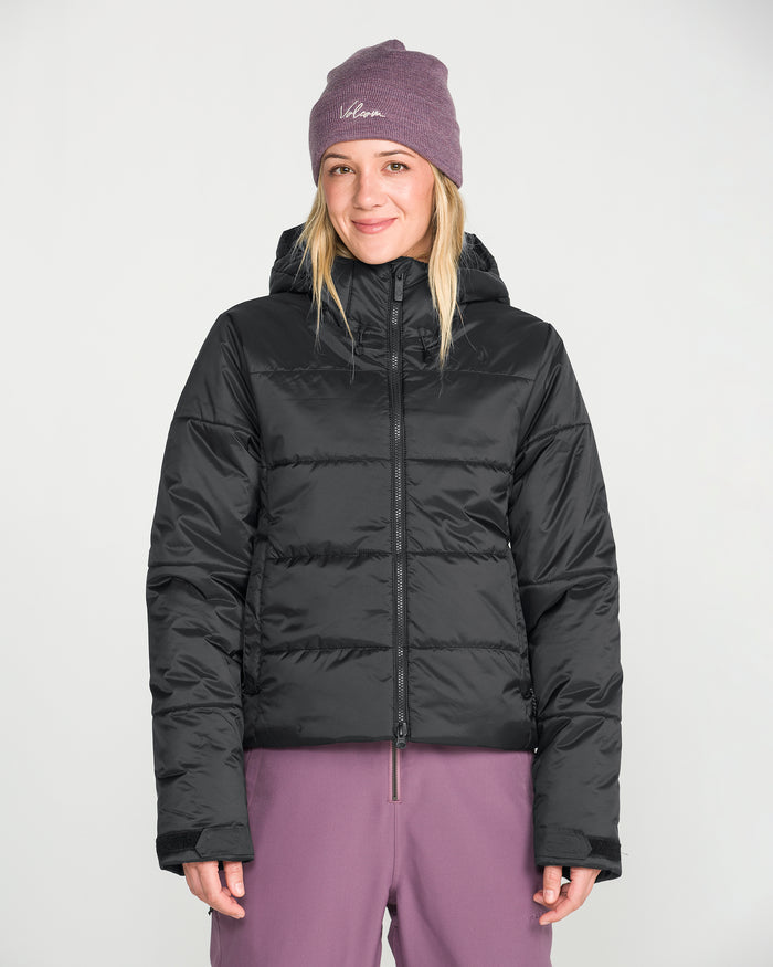 A person wearing a purple beanie, purple pants, and the Volcom Women’s Avalon Puff Jacket with recycled insulation stands facing forward against a plain white background, smiling slightly.