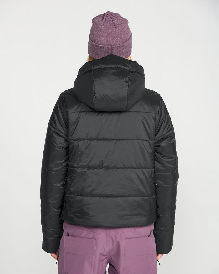 A person stands with their back to the camera, dressed in Volcom’s Women’s Avalon Puff Jacket—a black, waterproof jacket with recycled insulation—paired with mauve pants and a matching mauve beanie, set against a plain white background.