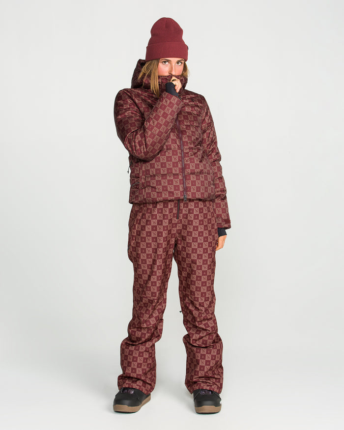 A person dressed in a maroon patterned winter ensemble stands against a plain white background, featuring the Volcom Women’s Avalon Puff Jacket with recycled insulation, matching pants and beanie, and black boots.