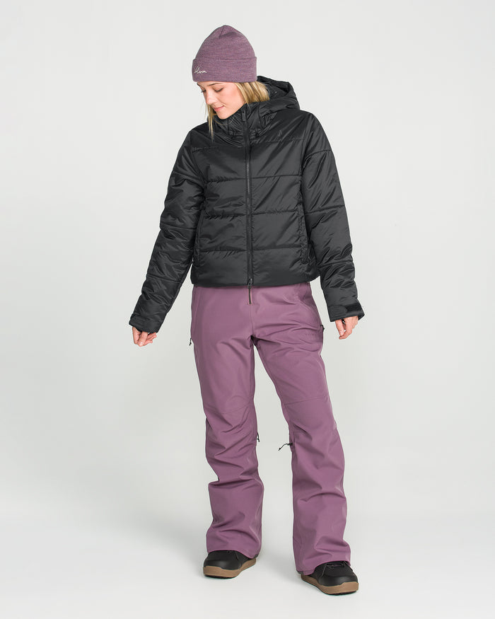 A person stands against a plain white background, looking down and to the side, wearing the Volcom Women’s Avalon Puff Jacket in black—a waterproof jacket featuring recycled insulation—paired with mauve snow pants and a mauve beanie.