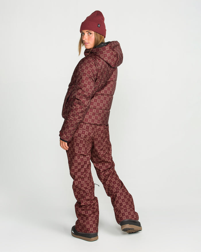 A woman models the Volcom Women’s Avalon Puff Jacket in a maroon pattern, paired with matching pants and a beanie, as she stands on a white background and looks over her shoulder.