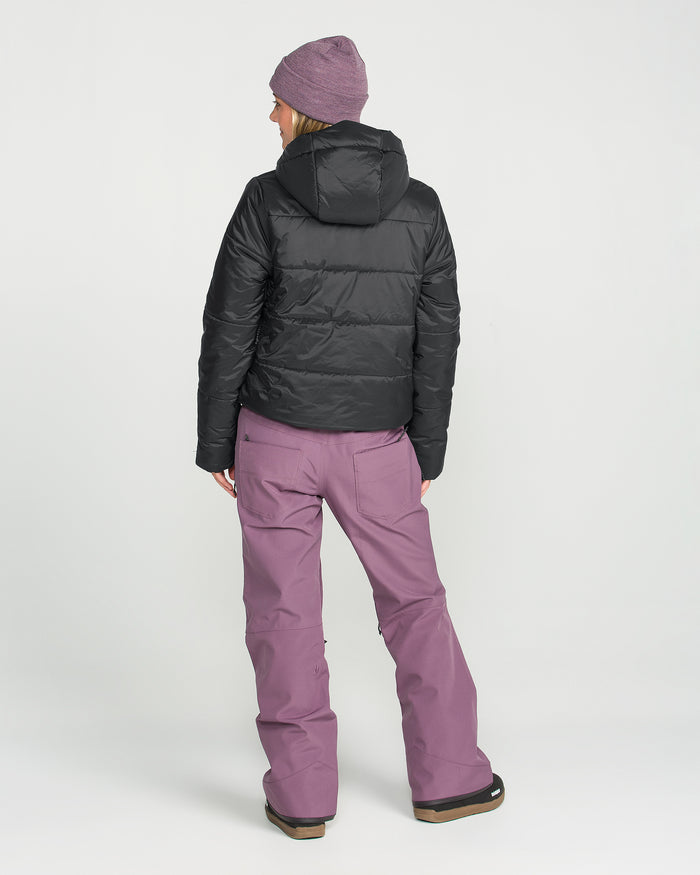 A person stands facing away, wearing the Volcom Women’s Avalon Puff Jacket—an insulated black hooded jacket—paired with a purple beanie and purple snow pants with pockets. The background is plain light gray.