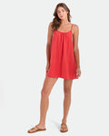 A woman wears the Volcom Women's Baby Cakes Mini, a sleeveless coral A-line dress, paired with brown sandals. She stands against a white background and looks at the camera with a relaxed expression.