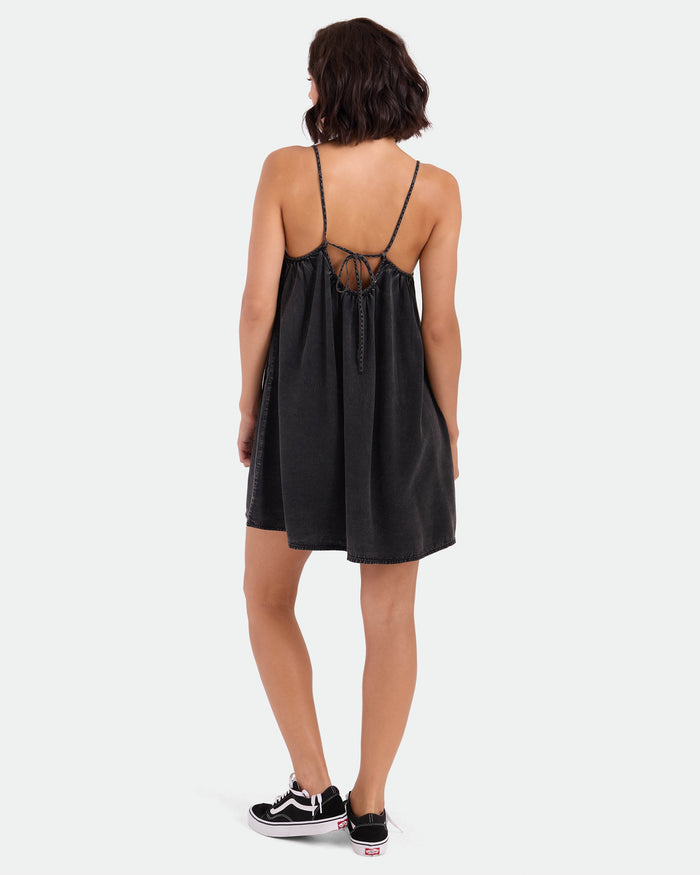 A person wears the Volcom Women's Baby Cakes Mini in Vintage Black—a loose, sleeveless woven day dress with adjustable tie-back and thin straps—paired with black and white sneakers, standing against a plain white background.