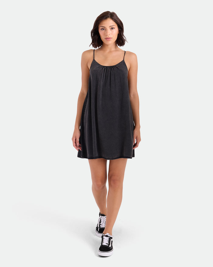 A woman with short brown hair wears the Volcom Women's Baby Cakes Mini, a vintage black woven day dress with thin straps and an adjustable tie-back, paired with black sneakers as she walks against a plain white background.