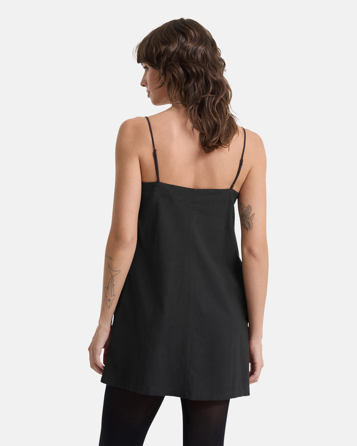 A woman with wavy brown hair and arm tattoos is seen from behind wearing the Volcom Women's Backbeatz Dress— a sleeveless black dress with thin straps— plus black tights, standing against a plain white background.