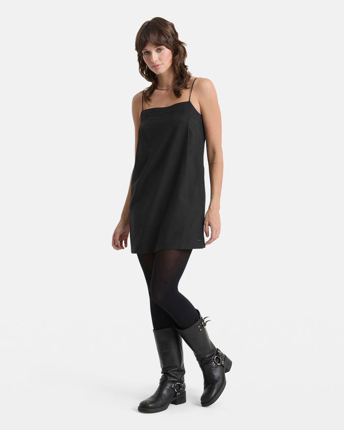 A woman with wavy brown hair poses casually against a white background wearing the Volcom Women's Backbeatz Dress, paired with black tights and buckled mid-calf boots.