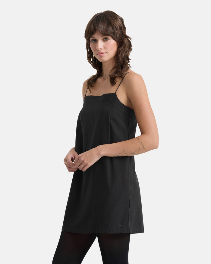 A woman with wavy brown hair models the Volcom Women's Backbeatz Dress, a sleeveless black above-the-knee style, standing against a plain white background with her hands loosely clasped and looking slightly to the side.