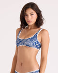 A woman with shoulder-length brown hair wears the Volcom Women's Bandana Scoop Bralette, featuring a blue and white pattern and crafted from recycled nylon. She stands against a plain, light background, looking at the camera with a neutral expression.
