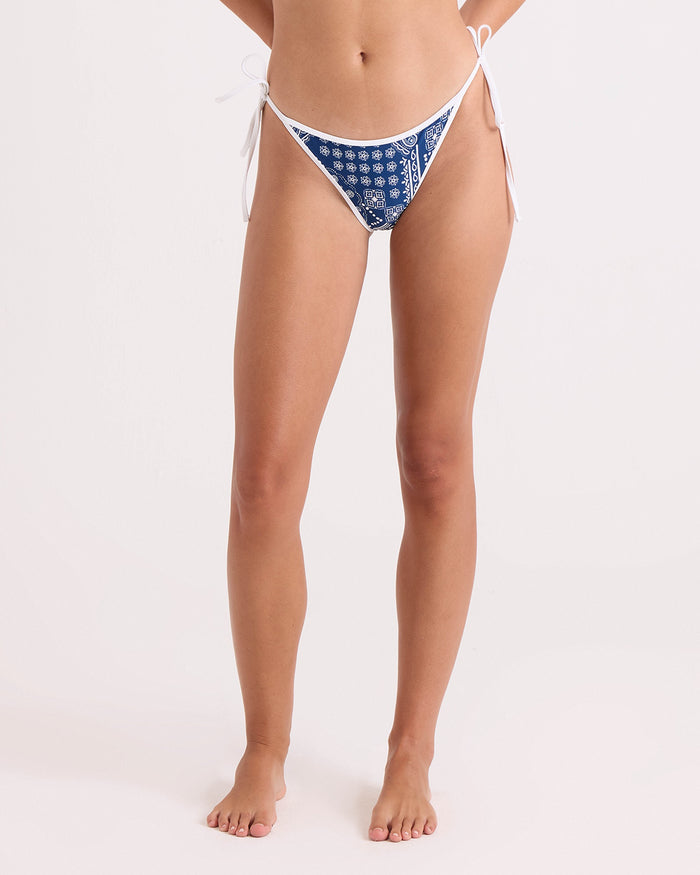 A woman stands barefoot against a plain light background wearing Volcom's Women's Bandana Tie Side Teeny Reversible bikini bottoms, featuring a blue and white pattern and crafted from recycled swimwear materials. Her head is not visible in the image.