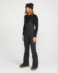 A woman wearing a black beanie, long-sleeve shirt, and Volcom Women’s Battle Stretch Bib—featuring PFAS-free DWR, 15K/10K protection, and stretch fabric—stands on a white background looking slightly to the side with a relaxed expression.