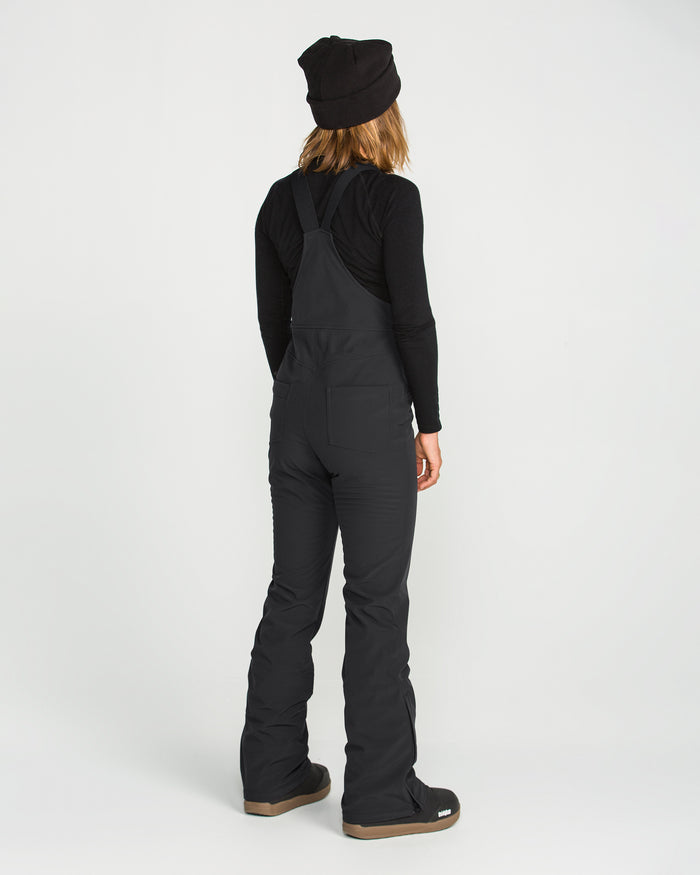 A person stands facing away from the camera against a plain white background, dressed in a black beanie, black long-sleeve shirt, and the Volcom Women’s Battle Stretch Bib in PFAS-free DWR black.