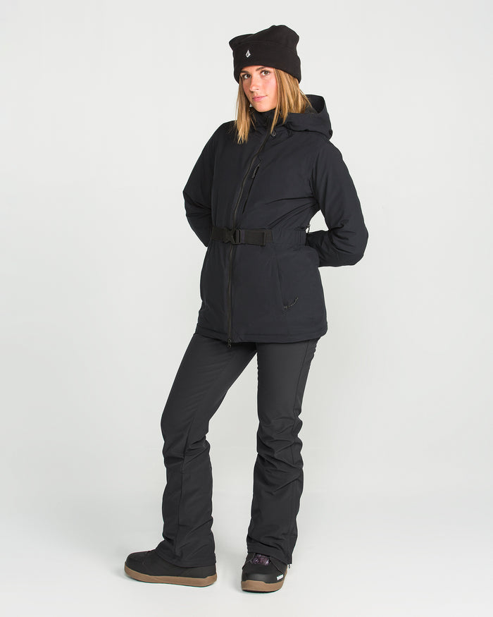 A person stands against a plain background wearing the Volcom Women’s Battle Stretch Bib paired with black boots and a black beanie, hands in pockets and striking a relaxed pose. The outfit features 15K/10K protection and PFAS-free DWR for enhanced weather resistance.