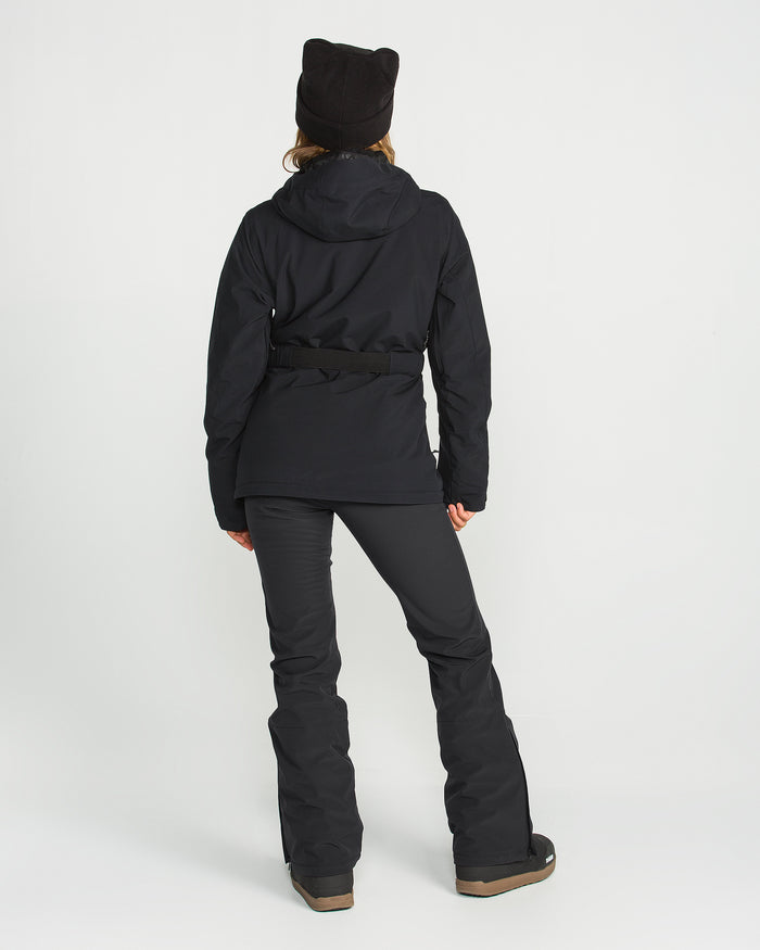 A person wearing the Volcom Women’s Battle Stretch Bib in black, paired with a black winter jacket featuring PFAS-free DWR, brown boots, and a black beanie stands facing away against a plain white background.