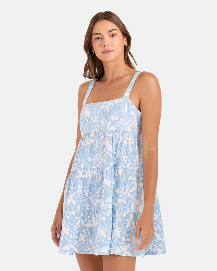 A woman models the Volcom Women's Beachmoonstone mini dress, featuring a light blue and white leaf print, as she stands against a plain background and looks at the camera with a relaxed expression.