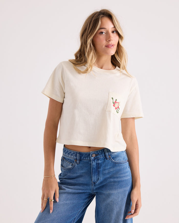 A woman wearing the Volcom Women's Bite Club Pocket Dial Tee—a cream-colored, 100% cotton cropped shirt featuring a floral design on the pocket—paired with blue jeans, stands against a plain light background.