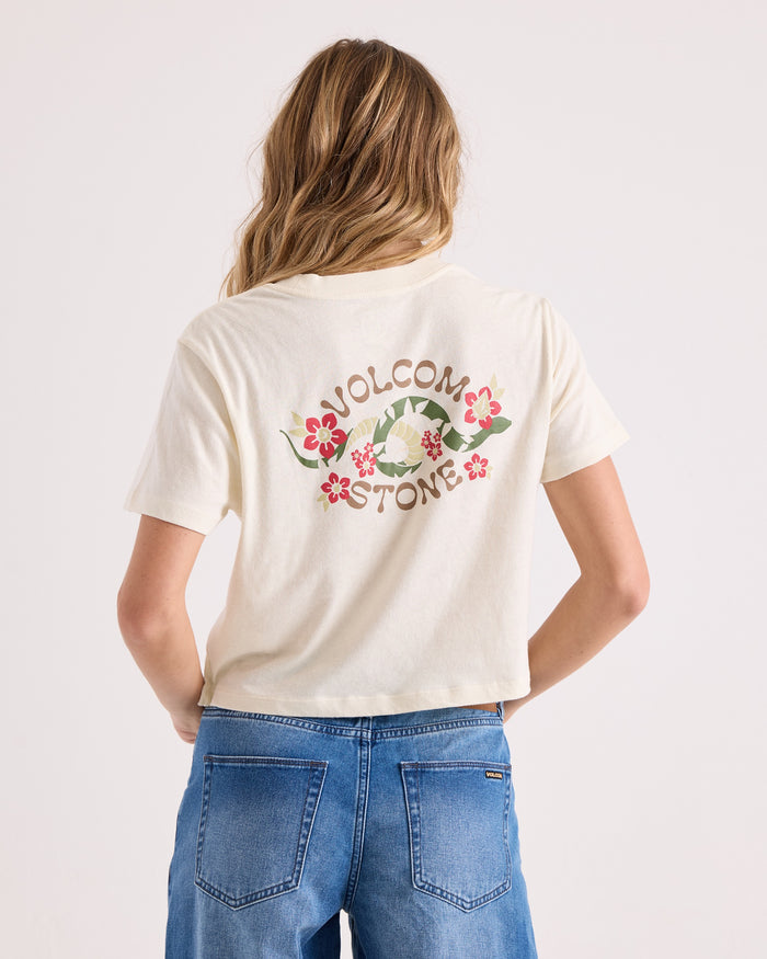 A person with long, wavy hair wears the Volcom Women's Bite Club Pocket Dial Tee—a white, cropped short-sleeve knit top made from 100% cotton, featuring Volcom Stone and flower graphics on the back—paired with blue jeans.