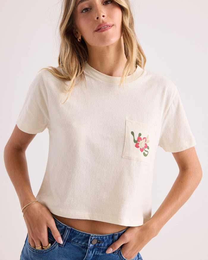 A woman is wearing the Volcom Women's Bite Club Pocket Dial Tee, a cream-colored short-sleeve knit top made from 100% cotton, featuring a flower design on the chest pocket. She pairs it with blue jeans and stands against a plain light background.