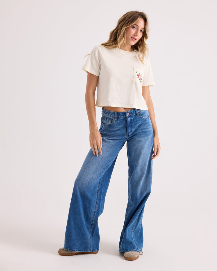 A person wearing the Volcom Women's Bite Club Pocket Dial Tee, a white 100% cotton cropped T-shirt with a small pocket design, paired with wide-leg blue jeans and brown shoes, stands posing against a plain light background.