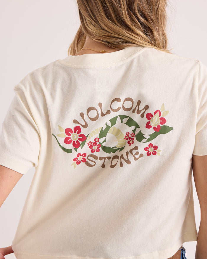 A woman wears the Volcom Women's Bite Club Pocket Dial Tee, a cream-colored short-sleeve knit top featuring "VOLCOM STONE" and floral graphics on the back. Only her back and partial arm are visible.