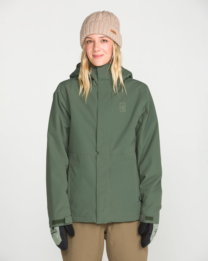 A woman stands against a plain white background, facing the camera and smiling slightly, dressed in a Volcom Women’s Bolt Insulated Jacket in green with V-Science 2-Layer technology, a light brown knit beanie, black gloves, and tan pants.