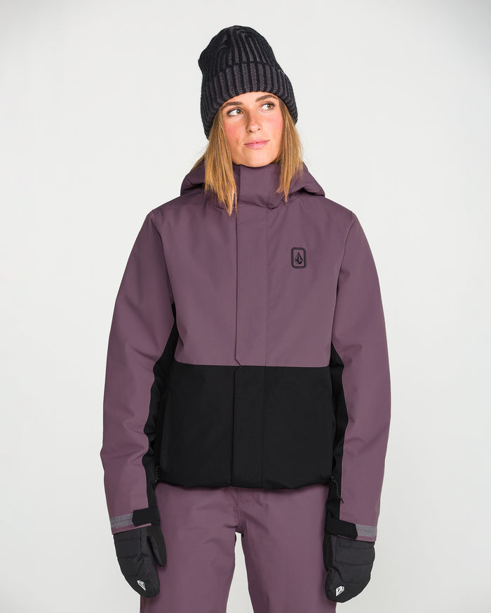 A person is wearing the Volcom Women’s Bolt Insulated Jacket in purple and black, paired with matching pants, black gloves, and a dark knit beanie, standing against a plain white background and looking slightly to the side.