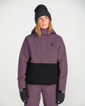 A person is wearing the Volcom Women’s Bolt Insulated Jacket in purple and black, paired with matching pants, black gloves, and a dark knit beanie, standing against a plain white background and looking slightly to the side.
