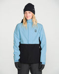 A person with long hair wearing a black beanie, black gloves, and dark pants stands in front of a plain light background, smiling slightly while modeling the Volcom Women’s Bolt Insulated Jacket—a light blue and black women’s jacket designed for recycled warmth.