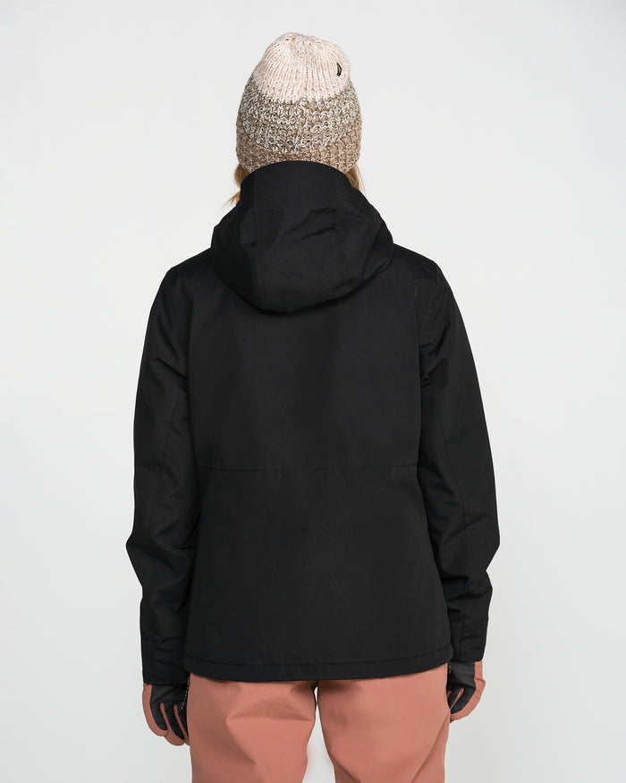 A person stands with their back to the camera against a plain white background, wearing the Volcom Women’s Bolt Insulated Jacket in black, a light knit beanie, and pink pants.