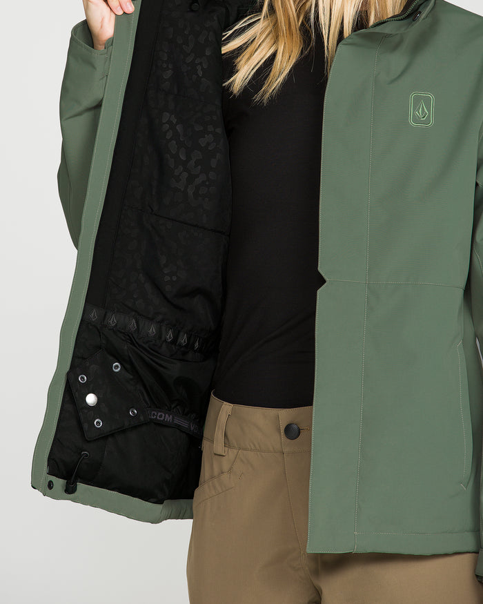 A person wearing the Volcom Women’s Bolt Insulated Jacket in green and brown pants opens the jacket to reveal its black, leopard-print lining, crafted with V-Science 2-Layer technology for recycled warmth. Only the lower half of their face and torso are visible, highlighting the sustainable style of this Volcom outerwear.