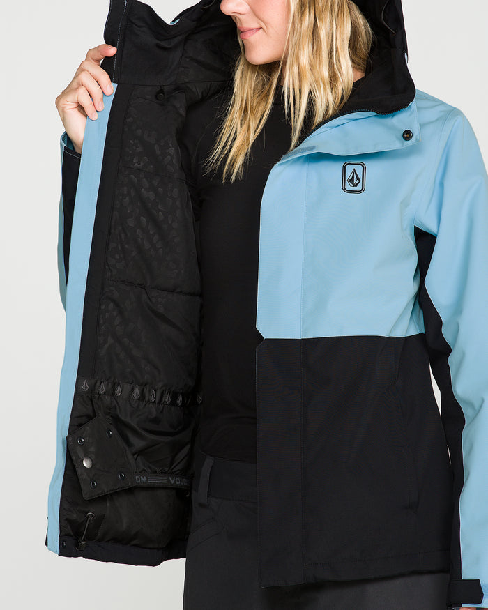 A woman holds open the Volcom Women’s Bolt Insulated Jacket in blue and black, displaying its inner lining, pockets, and snaps. With her face partially hidden by loose blonde hair, she emphasizes the jacket’s practical features—showcasing a design made for recycled warmth.