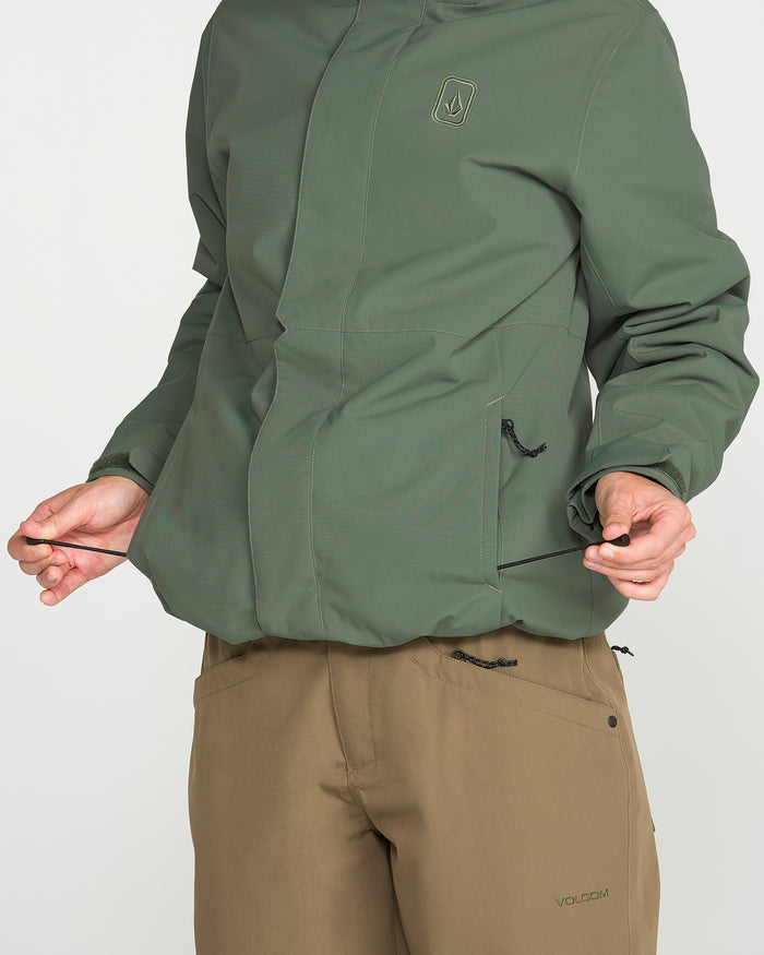 A person adjusts the drawstring on a green Volcom Women’s Bolt Insulated Jacket, featuring a small chest logo and flap-covered pockets for recycled warmth, paired with tan Volcom pants. The image is cropped below the neck.