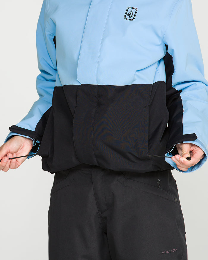 A person wearing the Volcom Women’s Bolt Insulated Jacket in light blue and black, along with black pants, pulls on the jacket’s waist drawstrings; only their torso and hands are visible.