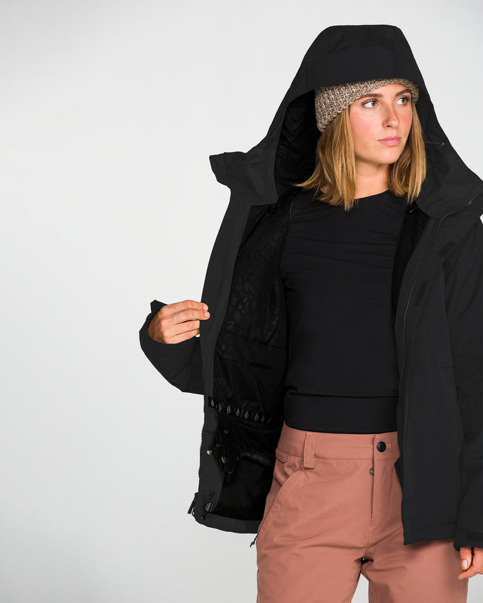 A person models the Volcom Women’s Bolt Insulated Jacket in black with the hood up, paired with a beige knit beanie, a black shirt, and pink pants, standing against a plain white background and looking to the side.