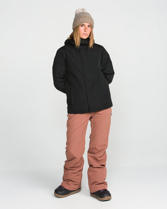 A person wearing a gray knit beanie, the Volcom Women’s Bolt Insulated Jacket in black, and loose-fitting pink snow pants stands against a plain white background with their hands behind their back.