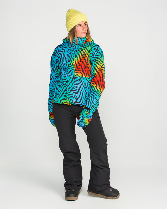 A person wearing a yellow beanie, black pants, and black boots stands against a plain white background, modeling the Volcom Women’s Bolt Insulated Jacket—a vibrant recycled warmth piece featuring colorful zebra stripes with blue, orange, green, and black accents.