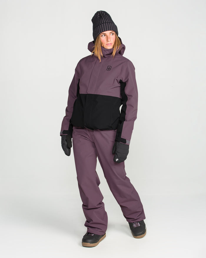 A person wearing the Volcom Women’s Bolt Insulated Jacket in black and purple, along with matching pants, black gloves, dark boots, and a dark beanie stands against a plain white background.