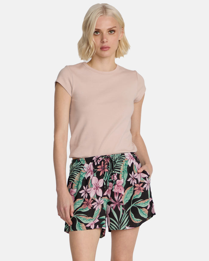 A woman with short blonde hair models Volcom's Women's Boss Beach Shorts, featuring a colorful pink and green tropical floral pattern, paired with a light pink short-sleeve top, against a plain white background.