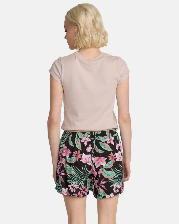 A woman with short blonde hair is seen from behind wearing a light pink short-sleeve shirt and Volcom Women's Boss Beach Shorts, which feature a black base with a vibrant pink and green floral design.