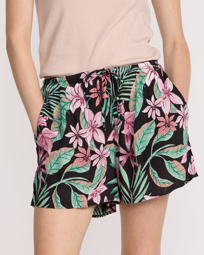 A person wears Volcom Women's Boss Beach Shorts featuring a vibrant pink and green tropical floral print, paired with a light pink top and hands in pockets.