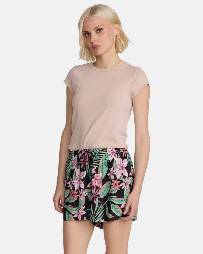 A woman with short blonde hair wears Volcom's Women's Boss Beach Shorts, featuring a colorful floral pattern, paired with a light pink short-sleeve top. She stands facing forward against a plain white background with a neutral expression.