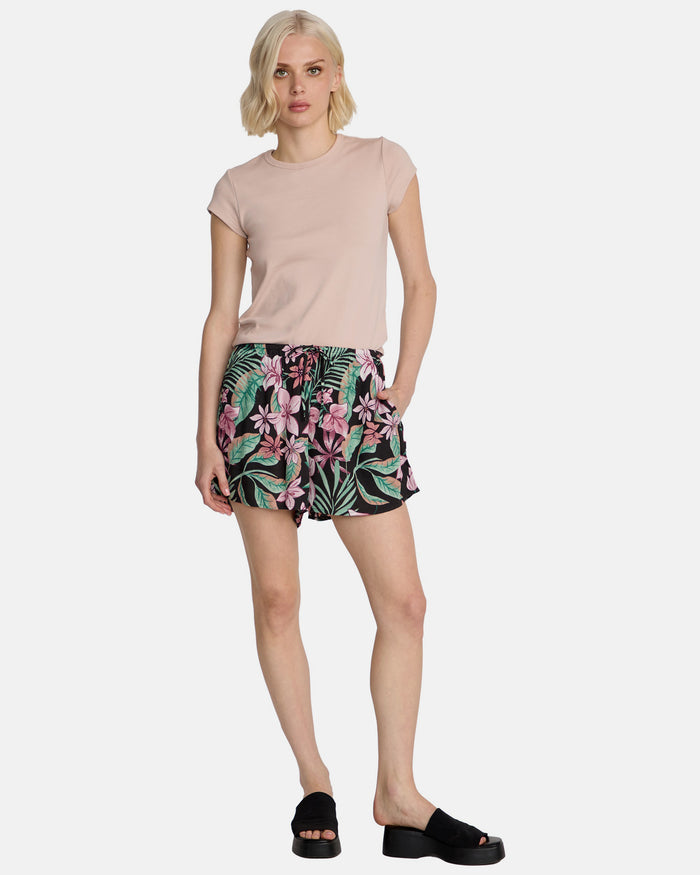 A woman with blonde hair wears Volcom Women's Boss Beach Shorts, a light pink t-shirt, and black slide sandals. She poses against a plain white background with one leg slightly bent.