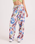 Wearing the Volcom Women's Bow Chicka Pants—loose, high-waisted, wide-leg trousers in a colorful tropical leaf and flower print with a relaxed fit and elastic waistband—paired with a light green crop top and white sneakers, the person stands against a plain background.