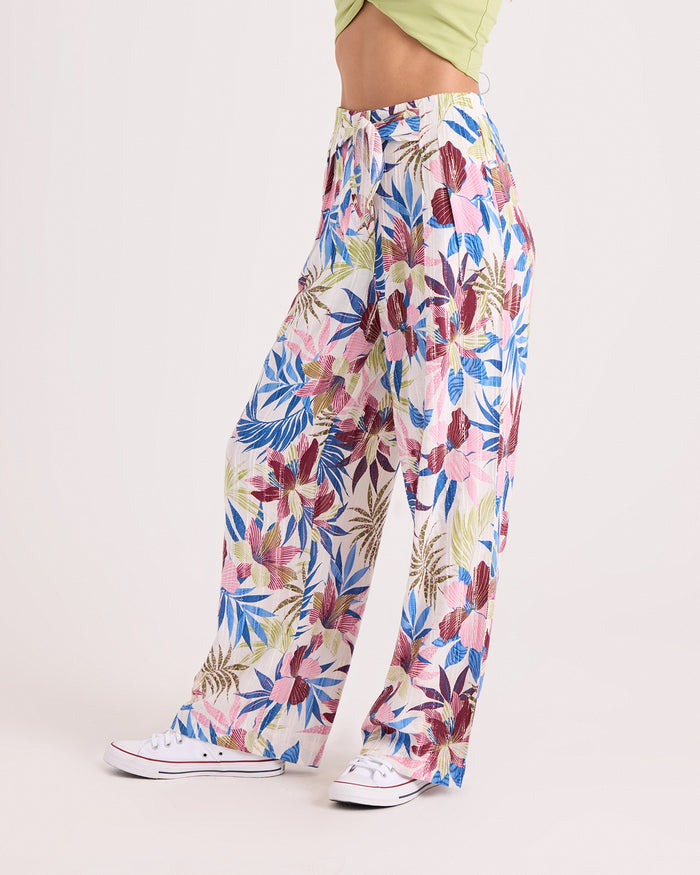 A person is wearing the Volcom Women's Bow Chicka Pants—relaxed fit, wide-leg, floral-patterned with an elastic waistband in blue, pink, and green—paired with white sneakers and a light green top, standing against a plain light background.
